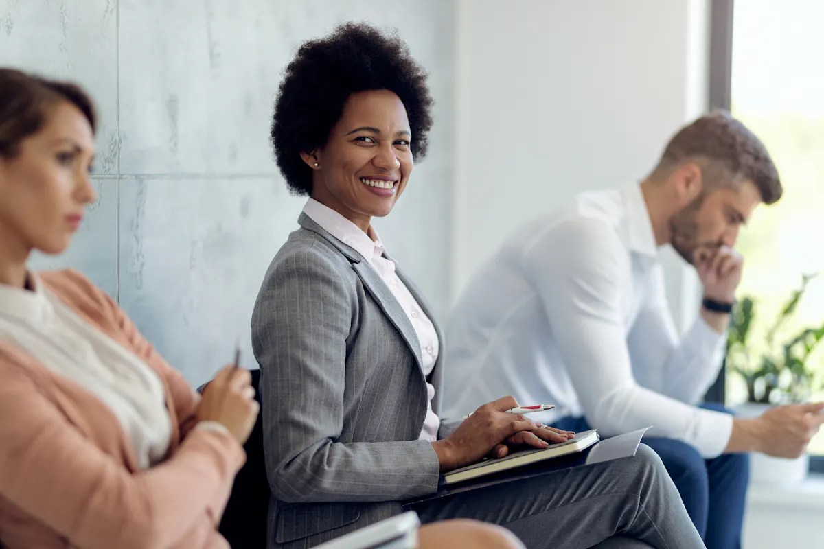 Three job candidates in business attire sit in a row with the middle individual sitting up straight and smiling.