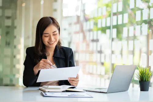 A professionally dressed person seated at a desk with an open laptop is reviewing information on a piece of paper.