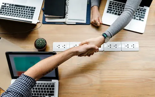 Two people grasp hands over a table with laptops on it