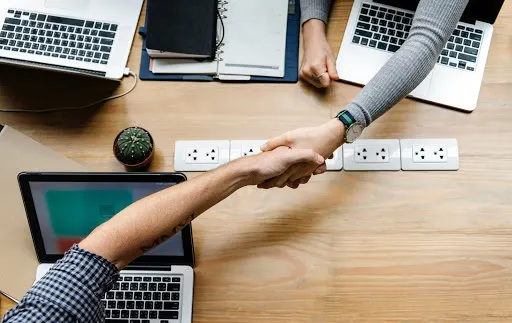 Two people grasp hands over a table with laptops on it